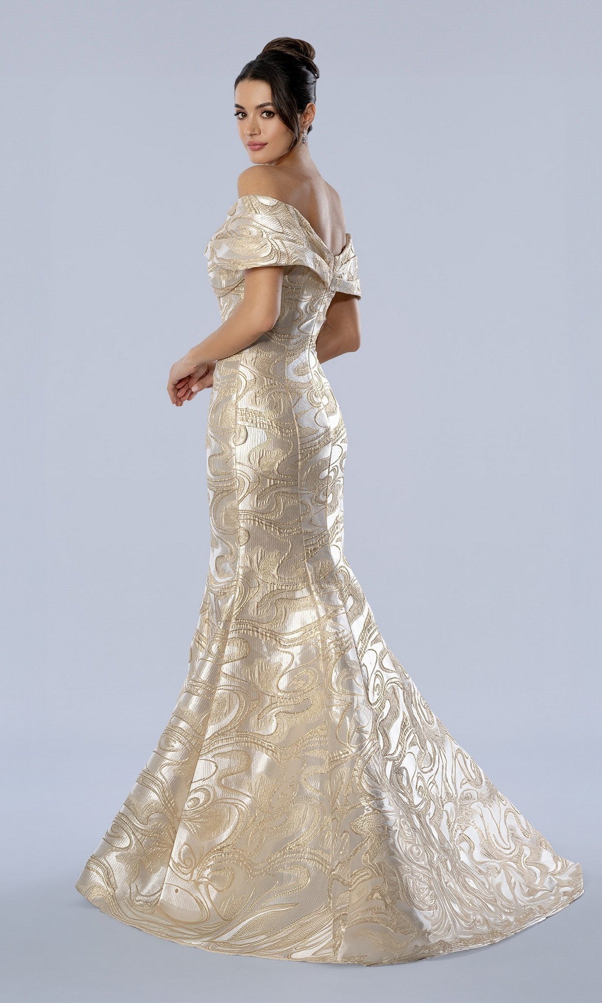 A woman poses against a plain background, looking over her shoulder in the Stella Couture 26360 long formal dress—an off-the-shoulder, floor-length gold gown adorned with an elegant swirl pattern reminiscent of metallic brocade.